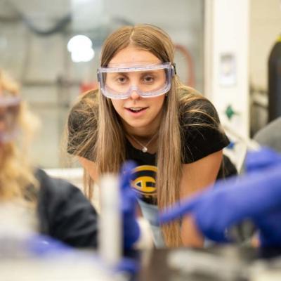 A student wearing safety goggles intently observing a laboratory experiment, with other students working alongside, focusing on the scientific process.